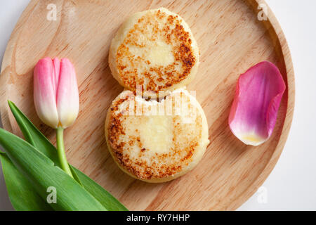 Nahaufnahme von frischen rosa Tulpe auf Holz- runde Platte, zwei Goldene gebissen fried Käsekuchen in der Form der Abbildung 8, Dessert von Hüttenkäse. Con Stockfoto