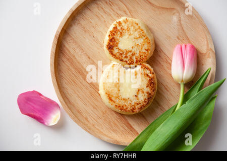 Nahaufnahme von frischen rosa Tulpe auf Holz- runde Platte, zwei Goldene gebissen fried Käsekuchen in der Form der Abbildung 8, Dessert von Hüttenkäse. Con Stockfoto