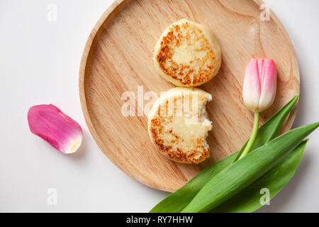 Nahaufnahme von frischen rosa Tulpe auf Holz- runde Platte, zwei Goldene gebissen fried Käsekuchen in der Form der Abbildung 8, Dessert von Hüttenkäse. Con Stockfoto