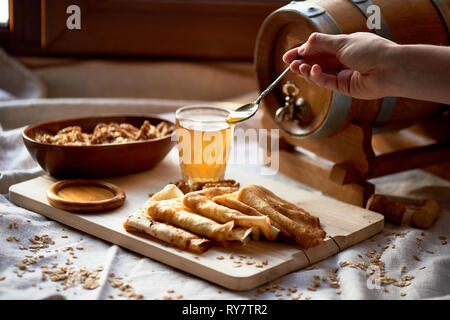 Mead und Pfannkuchen. Russische Gericht für Faschingsdienstag. Abschied vom Winter, die Hand in den Rahmen. Honig vom Löffel tropft auf Pfannkuchen Stockfoto