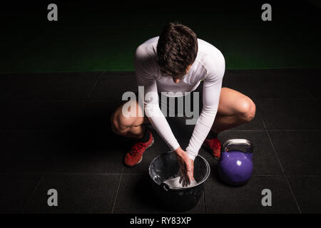 Von oben athletischen jungen Kerl in Sportkleidung kreidung Hände zum Heben von großen kettlebell in der Turnhalle Stockfoto