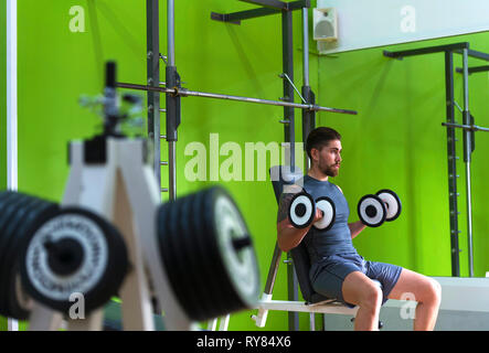 Sicher mann Hanteln während auf Sitz gegen grüne Wand im Fitnessraum Stockfoto