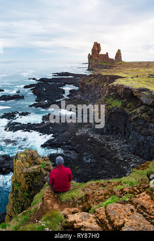 Tourist guy sitzt auf einem Felsen und blickt auf das Meer. Die Klippen von Londrangar, einer berühmten Touristenattraktion von Island Stockfoto