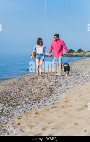 Ehepaar mit Hund, das Hände beim Gehen am Strand gegen den blauen Himmel während der sonnigen Tag Stockfoto