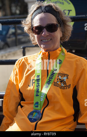 Frau (von Hove Geländeläufer) mit Goldmedaille für den Abschluss von Brighton Halbmarathon am 24. Februar 2019, entspannend auf Wand an der Seite der Republik Griechenland Gre Stockfoto