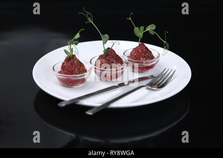 Rote schmackhafte Sauce Tartar in drei pialas auf einem Teller mit Messer und Gabel, mit einem Schaft der Gekeimten grüne Erbsen eingerichtet, Nahaufnahme, für ein Restaurant Stockfoto