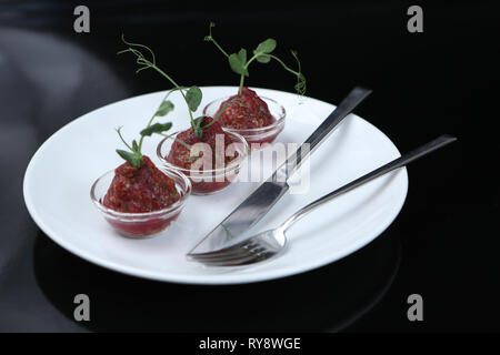 Rote schmackhafte Sauce Tartar in drei pialas auf einem Teller mit Messer und Gabel, mit einem Schaft der Gekeimten grüne Erbsen eingerichtet, Nahaufnahme, für ein Restaurant Stockfoto