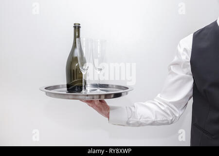 Ein Kellner/Butler in weißem Hemd und schwarzen Anzug Weste mit einem silbernen Tablett mit einer Flasche Champagner und zwei Gläser. Weißer Hintergrund. Stockfoto