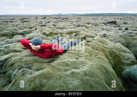 Mann Reisender in roter Jacke liegen auf dem Moos. Lavafeld an der südlichen Küste von Island, Europa. Touristische Attraktion. Erstaunlich in der Natur. Bewölkt Sommer d Stockfoto