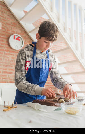 Low Angle View der Boy verzieren Schokolade Kuchen auf dem Tisch, während gegen die Mauer zu Hause stehend Stockfoto