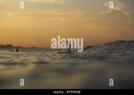 Seitenansicht der Silhouetten der Männer mit Paddle Surf Board zwischen Wasser von Himmel und Meer in Abend auf Bali, Indonesien Stockfoto