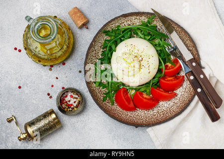 Gourmet Italienisch Käse Burrata (Mozzarella und strachatella) auf einem braunen Ton Platte mit frischem Rucola, Tomaten, frisch gemahlenem Pfeffer und ol serviert. Stockfoto