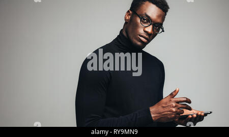 Afrikanische Mann in Schwarz Polo-T-Shirt mit seinem Smart Phone und Kamera. Hübscher Kerl mit einem Mobiltelefon auf grauem Hintergrund. Stockfoto