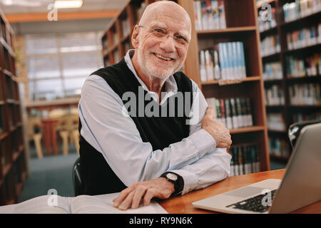 Lächelnd älterer Mann sitzt in einer Bibliothek mit seinen Büchern und Laptop auf dem Tisch. Fröhlicher älterer Mann in Bibliothek und lernen sitzen. Stockfoto