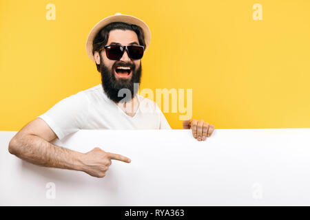 Studio Portrait eines bärtigen Kerl in der Sonnenbrille und Hut, mit Blick hinter die leeren Banner und weist ihn an, Bild auf gelbem Hintergrund Stockfoto