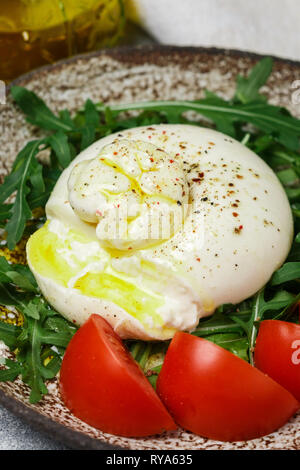 In Scheiben geschnitten Gourmet Italienisch Käse burrata serviert auf einem braunen Ton Platte mit frischem Rucola, Tomaten, frisch gemahlenem Pfeffer und Olivenöl mit Gabel und k Stockfoto