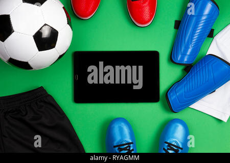 Fußball-Fußball-Hintergrund. Tablet Computer mit Fußball, blau Stiefel, Klampen, weißes T-Shirt und schwarze Shorts auf grünem Hintergrund. Ansicht von oben Stockfoto