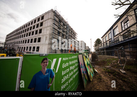 Wiesbaden, Deutschland. 12 Mär, 2019. Poster auf dem Bauzaun über das neue Gebäude des "Dr.-Horst-Schmidt-Klinik' (l Informieren). Der Betreiber, Helios Verwaltung Hessen GmbH, beabsichtigt, das Gebäude bis 2021 abgeschlossen und wird 268 Millionen Euro investieren. Die alte Klinik Gebäude (r) ist von 2023 abgerissen werden. Credit: Andreas Arnold/dpa/Alamy leben Nachrichten Stockfoto