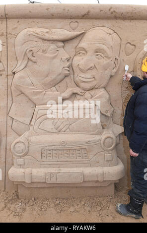 Binz, Deutschland. 12 Mär, 2019. Der Bildhauer Wlodek Bludnik aus Polen auf dem Sand Skulptur 'Berliner Mauer' arbeitet mit einer modernen Version des graffito der Bruder Kuss auf dem Gelände der Sandskulpturenfestival. Es zeigt uns Präsident Donald Trump und der russische Präsident Wladimir Putin. Künstlerinnen und Künstler aus verschiedenen europäischen Ländern werden derzeit erstellen 45 riesige Zahlen für die 10 Sand Skulptur zeigen im Ostseebad Binz. Der Sand Skulpturen zeigen, eröffnet am 16.03.2018. Quelle: Stefan Sauer/dpa-Zentralbild/dpa/Alamy leben Nachrichten Stockfoto