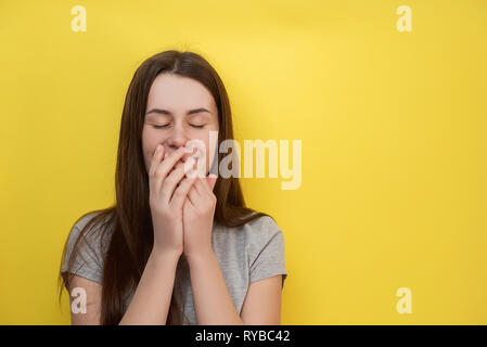 Horizontale Schuß von gut aussehenden jungen Mädchen freudig kichert, deckt den Mund so versucht zu Lachen, trägt graue t-shirt auf gelbe Wand isoliert. Menschen ein Stockfoto