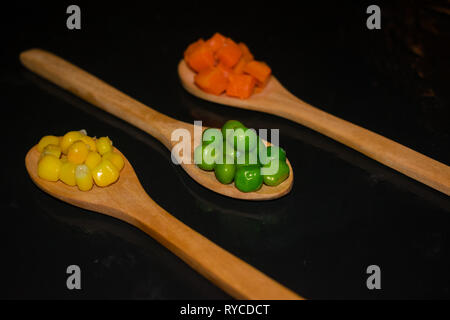 Stücke von Karotten, mais und Erbsen auf Holzlöffel mit dunklen Hintergrund Stockfoto