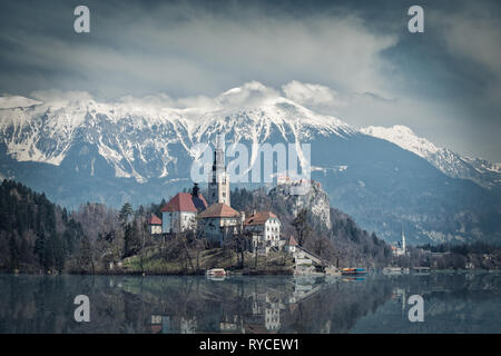 Bled Kirche auf einer Insel im Wasser der See mit Snowy Mountain Range Karawanks und Berg Hochstuhl Stol auf einem sehr bewölkten Tag im Frühjahr Bled reflektierte ich Stockfoto