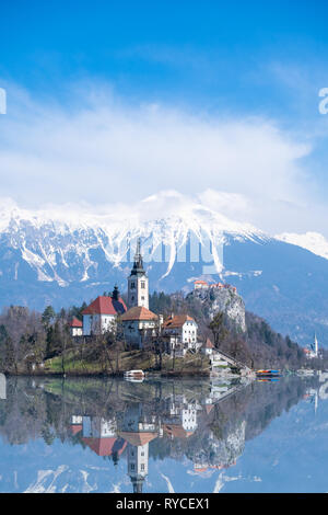 Bled Kirche auf einer Insel im Wasser der See mit Snowy Mountain Range Karawanks und Berg Hochstuhl Stol an einem bewölkten Tag im Frühling in Slo Bled wider Stockfoto