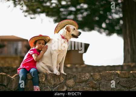 Junges Mädchen sitzt auf einer Wand mit ihren Arm um einen Hund, beide tragen Cowboyhüte. Stockfoto