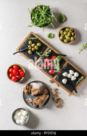 Mozzarella, Tomaten, Oliven, Antipasti Vorspeisen in Schüsseln und auf Schiefer, das Board mit Basilikum, Rucola und Roggenbrot über grau Textur serviert. Stockfoto