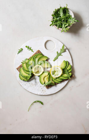 Veganen Sandwiches mit in Scheiben geschnittenen Avocado und Zitrone auf Roggenbrot, Rucola Salat auf Keramik Brett über weißem Marmor Hintergrund serviert. Flach, Raum Stockfoto