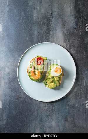 Vegetarisches Sandwich mit in Scheiben geschnittenen Avocado, sonnengetrocknete Tomaten, Ei, Garnelen, auf keramische Platte über blaue Textur Hintergrund serviert. Flach, Raum Stockfoto