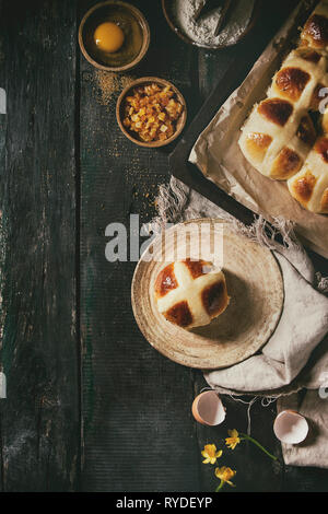 Hausgemachte Ostern traditionelle Hot Cross Buns auf Platte und ein Backblech mit Backpapier auslegen und Zutaten oben auf dunklem Hintergrund. Ansicht von oben, Raum Stockfoto