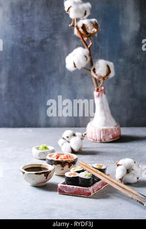 Sushi Rollen in rosa Keramik Servierteller mit Stäbchen, Schüsseln aus Sojasauce, und eingelegtem Ingwer, Baumwolle Blumen auf hellblau Tabelle. Japan Menü. Stockfoto