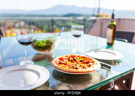 Frische hausgemachte Pizza auf Glas Tisch auf der Terrasse draußen in Italien mit Tomatensauce, Gemüse und zwei leeren weißen Teller und Gläser Rotwein Grün Stockfoto