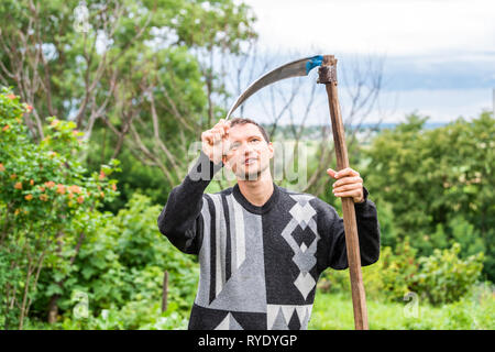 Glückliche junge Mann Bauer im Garten stehen mit Sichel Sense rake Werkzeug in grün sommer in der Ukraine durch landwirtschaftliche Pflanzen Stockfoto