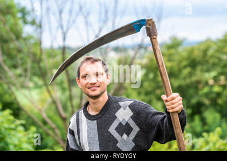 Glückliche junge Mann Nahaufnahme Gesicht Bauer im Garten stehen mit Sichel Sense rake Werkzeug in grün sommer in der Ukraine durch landwirtschaftliche Pflanzen Stockfoto