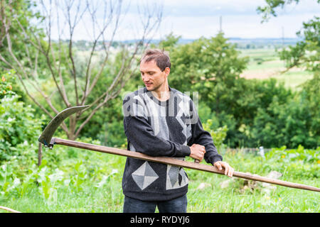 Junger Mann Bauer im Garten stehen mit Sichel sense Tool in grün sommer in der Ukraine durch landwirtschaftliche Pflanzen Stockfoto