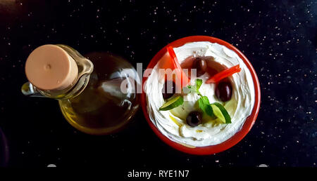 Eine Schüssel Labneh, ein arabischer Joghurt-Frischkäse-Dip, mit Gemüse und einer Flasche Olivenöl auf einem Tisch. Küche des Nahen Ostens. Stockfoto