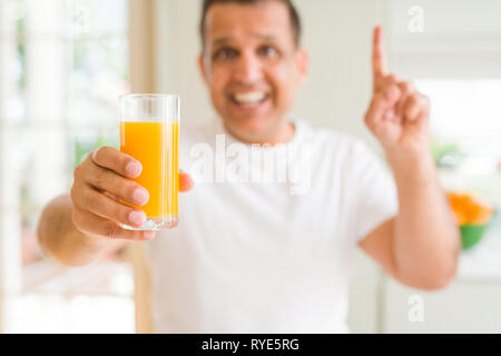 Mittleres Alter Mann trinkt ein Glas Orangensaft zu Hause mit einer Idee oder Frage Zeigefinger mit glücklichen Gesicht überrascht, Nummer eins Stockfoto