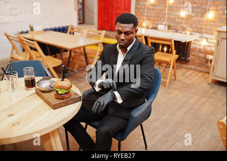 Das Tragen von Handschuhen für Lebensmittel. Ehrbare junge afrikanische amerikanische Mann im schwarzen Anzug sitzt im Restaurant mit leckeren double Burger. Stockfoto