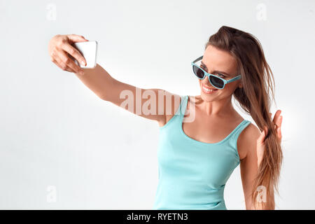 Wundervolle Momente. Frau in Sonnenbrille ständigen Studio isoliert auf weißem Unter selfie auf dem Smartphone berühren Haar verspielter Stockfoto
