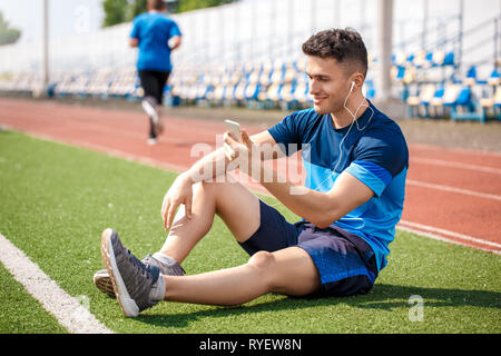 Junger Mann in Ohrhörer im Stadion sitzen die Wiedergabe von Videos auf dem Smartphone fröhliche Seite anzeigen Stockfoto