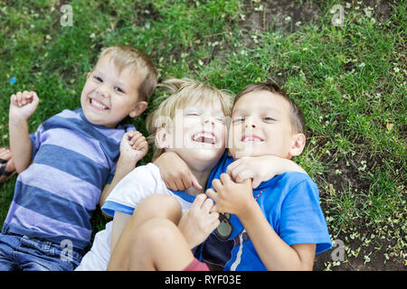 Drei glückliche Jungs im Sommer Park. Freunde oder Geschwister umarmen und Lachen im Liegen auf grünem Gras. Stockfoto