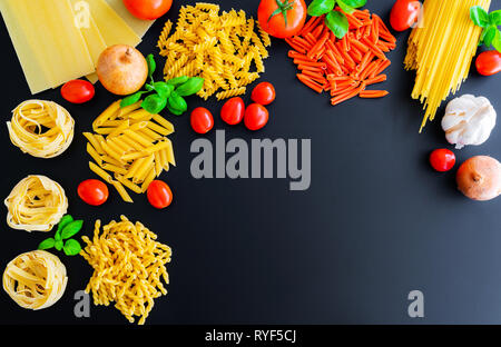 Verschiedene ungekochte italienische pasta Nudeln auf dunklem Hintergrund mit Basilikumblätter, frischen Tomaten und Zwiebeln Stockfoto
