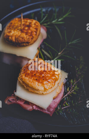 Vertikale Foto mit zwei Sandwiches. Sandwiches sind aus Blätterteig Teig Quadrate mit Käse gefüllt und getrocknetem Schinken. Essen ist auf schwarze Platte mit platziert Stockfoto