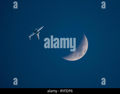 Eines niedrig fliegenden Verkehrsflugzeug passiert einen Halbmond in einem dunkelblauen Himmel bei Sonnenuntergang. Stockfoto