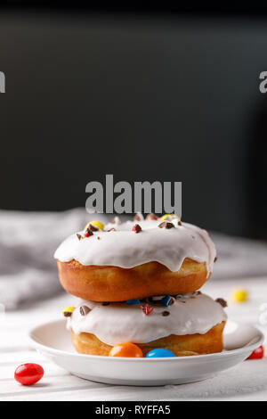 Kochen, Backen und Essen Konzept - Nahaufnahme. Zwei Donuts in weißer Glasur mit farbigen Schokolade Dragiermaschinen u.a. auf einem Teller. Noch appetitlich Leben. Stockfoto