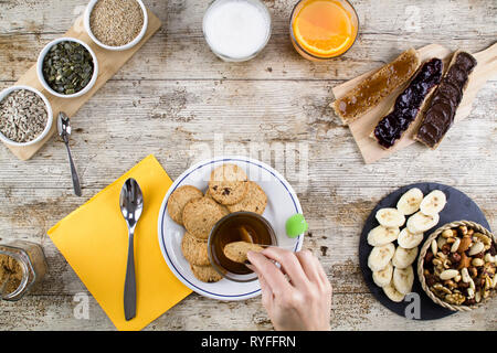 Eine Frau hand Einweichen der Keks in Kaffee an einem Holztisch zum Süßen Veganes Frühstück Schuß von oben mit Bananenscheiben, gemischte getrocknete Früchte, Gemüse Stockfoto