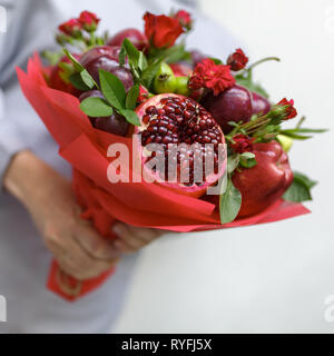 Genießbare Blumenstrauß aus Granatapfel, Äpfel, Pflaumen und roten Rosen in die Hände der Frau auf weißem Hintergrund Stockfoto