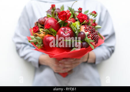 Genießbare Blumenstrauß aus Granatapfel, Äpfel, Pflaumen und roten Rosen in die Hände der Frau auf weißem Hintergrund Stockfoto